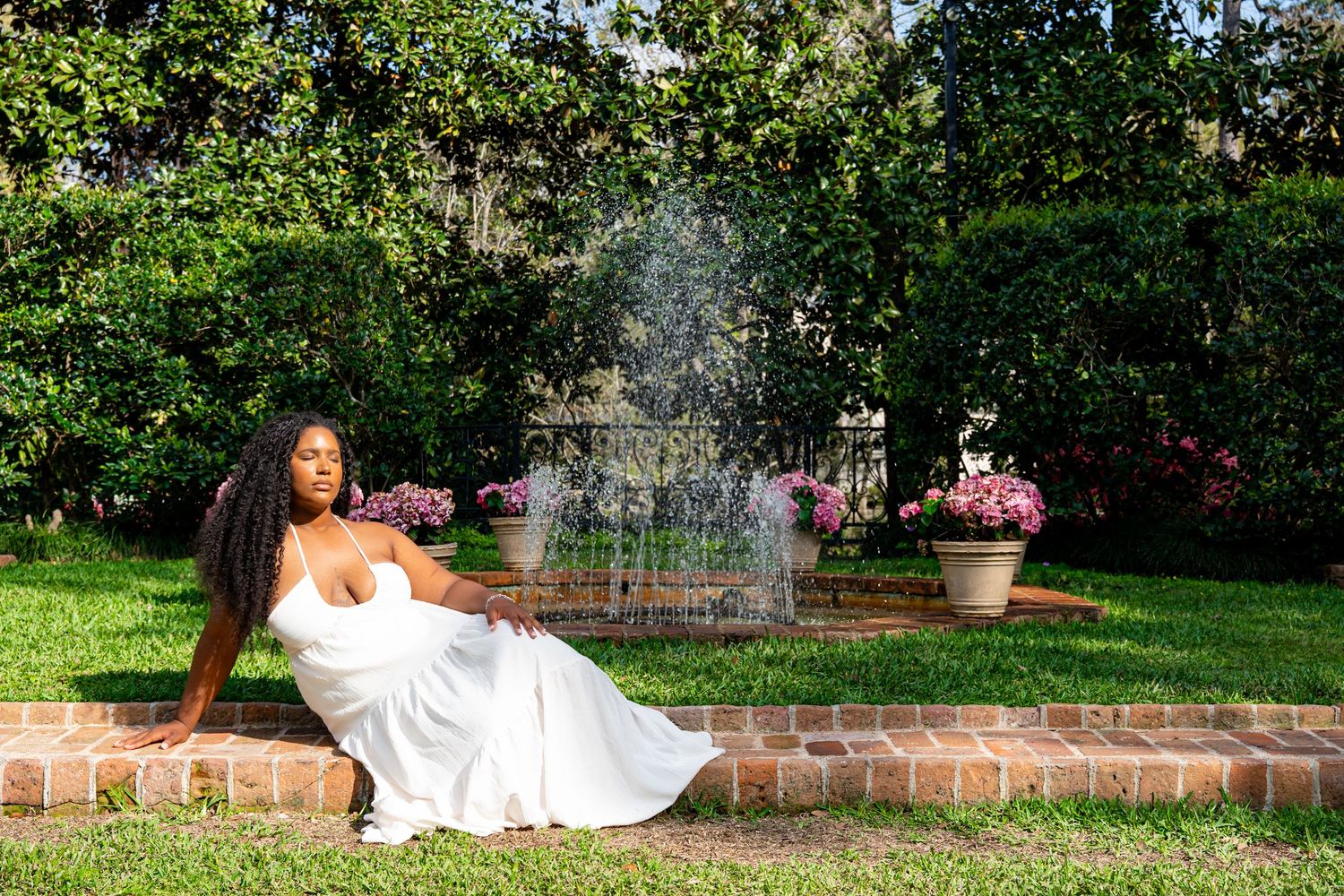 Woman in white maxi dress sitting by a garden fountain surrounded by lush greenery and flowers.