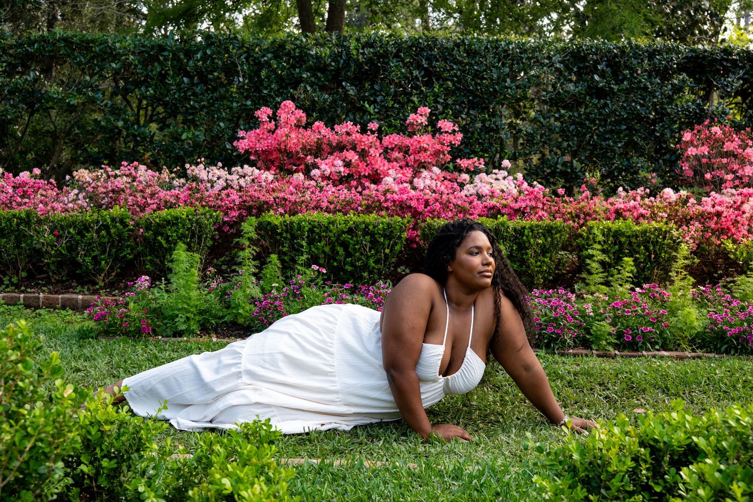 Woman in white dress posing on grass before a hedge wall of vibrant pink blooming roses.