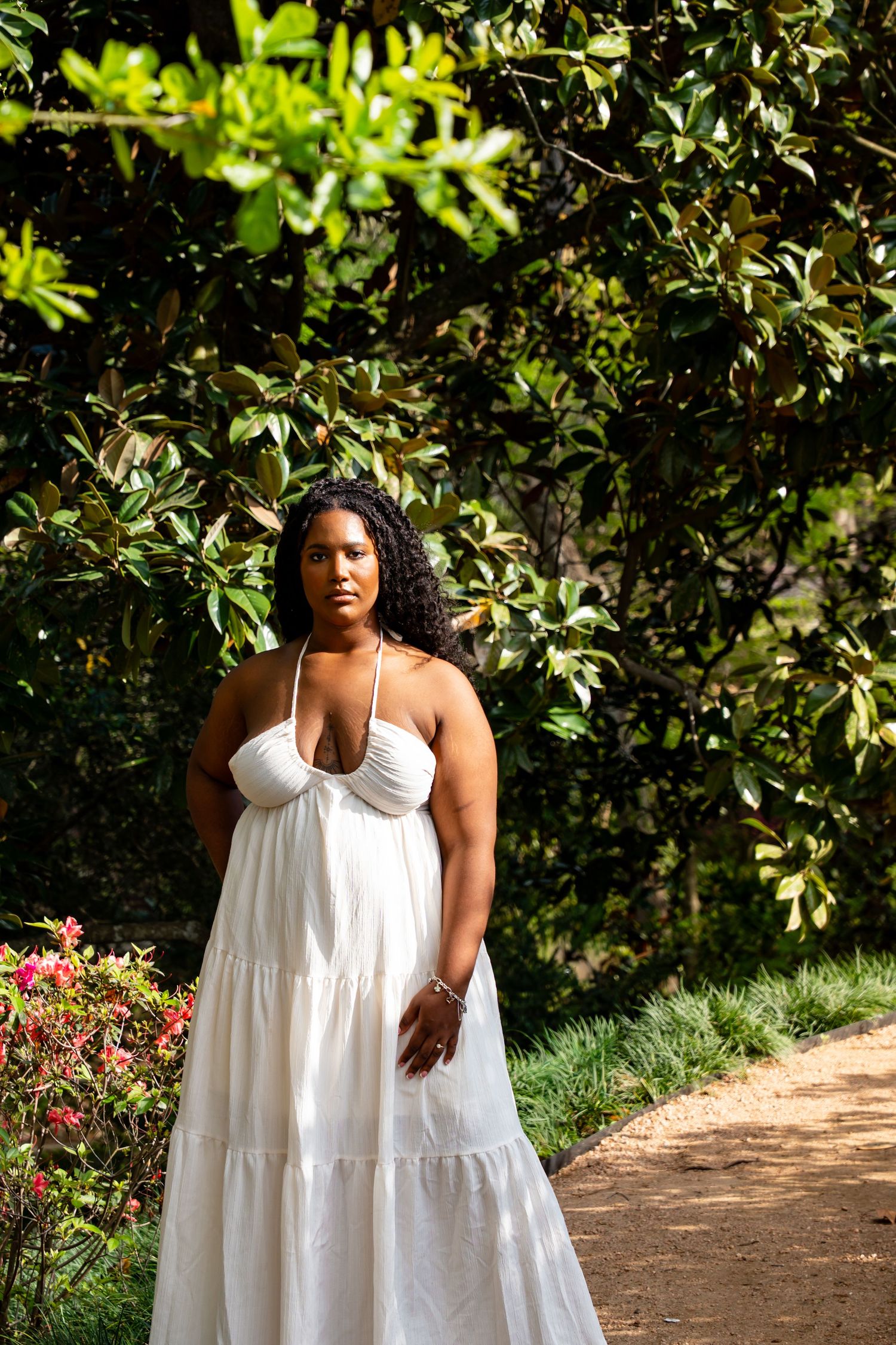 Woman in white halter maxi dress standing on a garden path beneath a large magnolia tree.