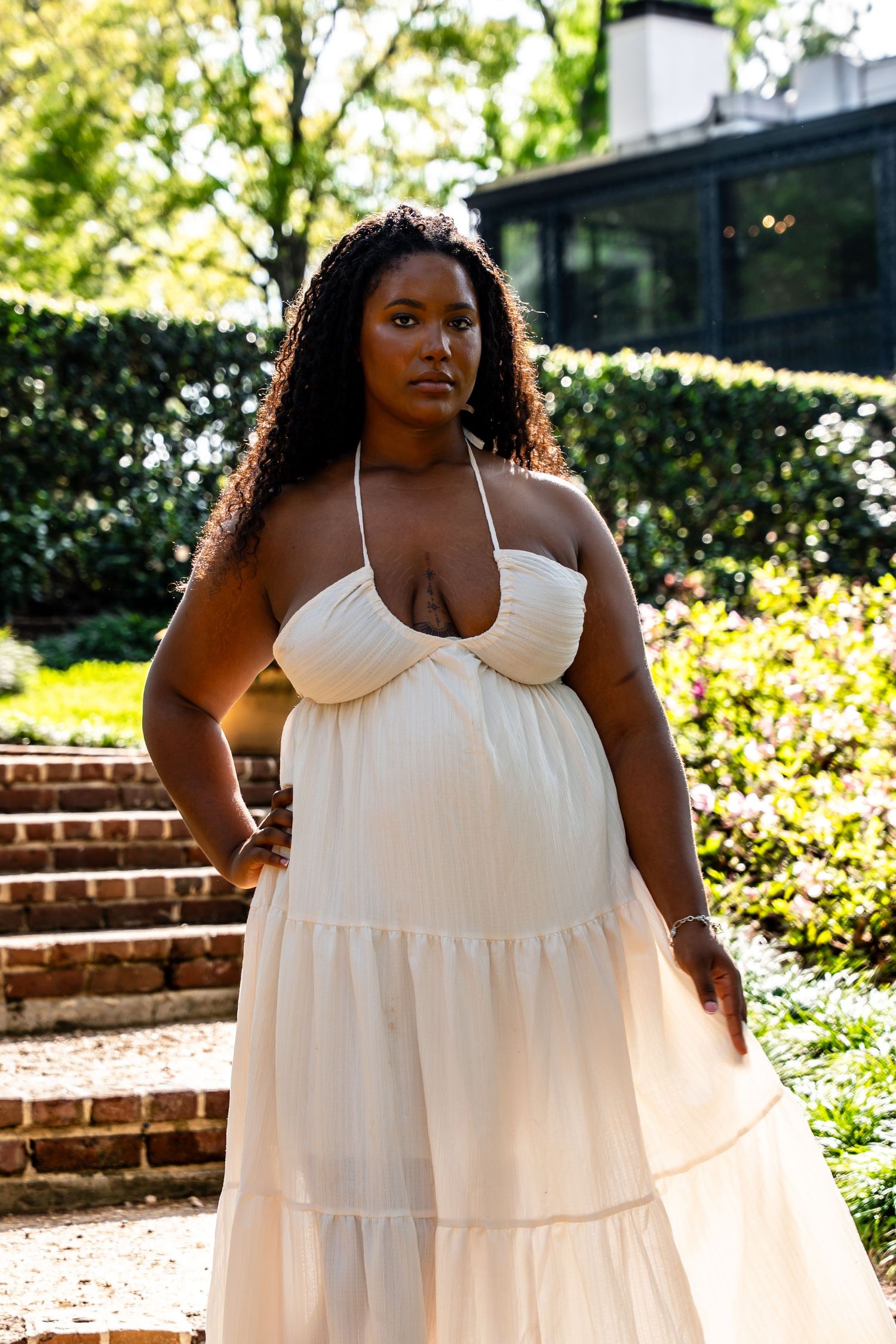 Woman in cream halter maxi dress posing outdoors near brick steps in a sunlit garden setting.