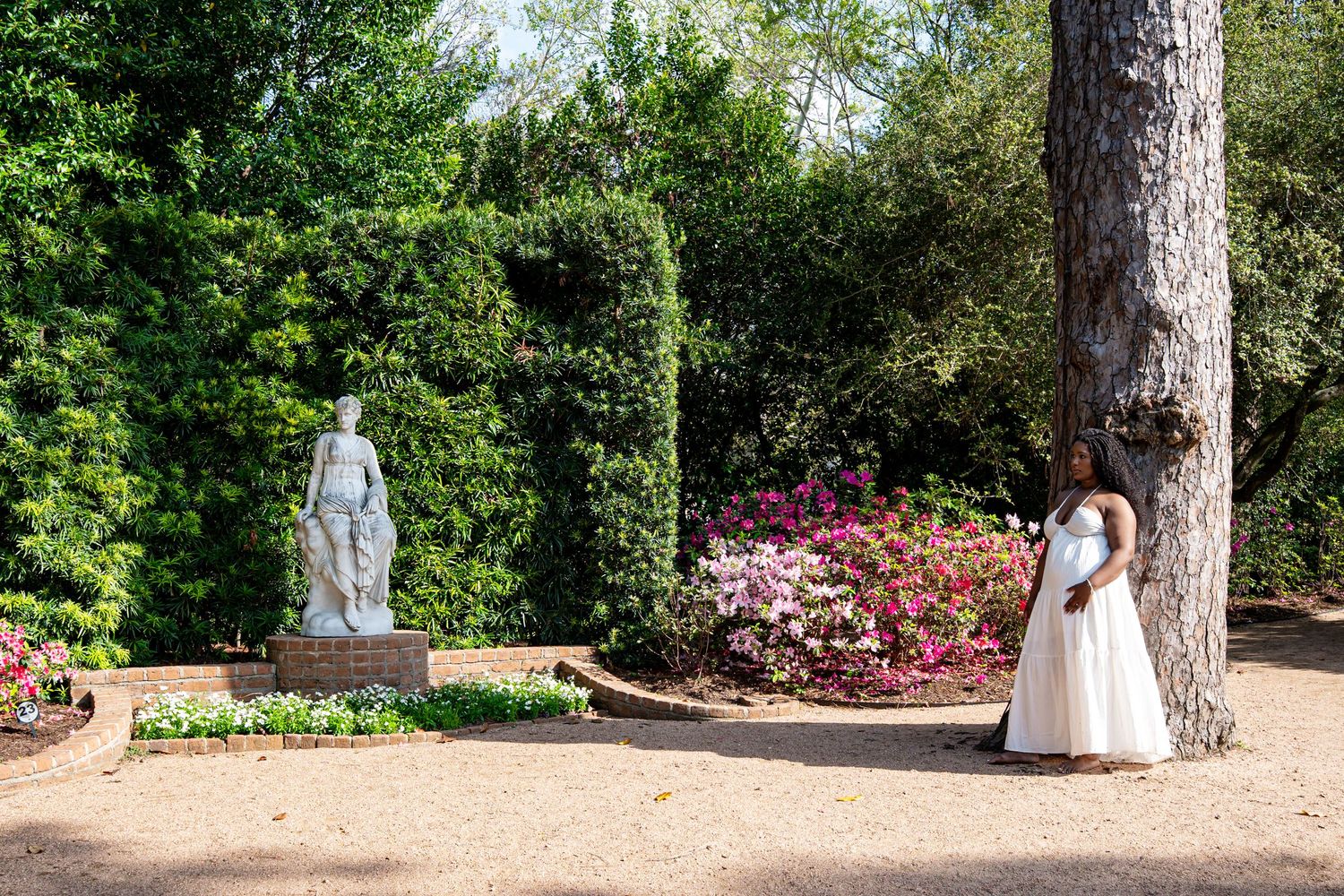 Woman in white dress standing by a large tree near a classical garden statue amid pink flowers.