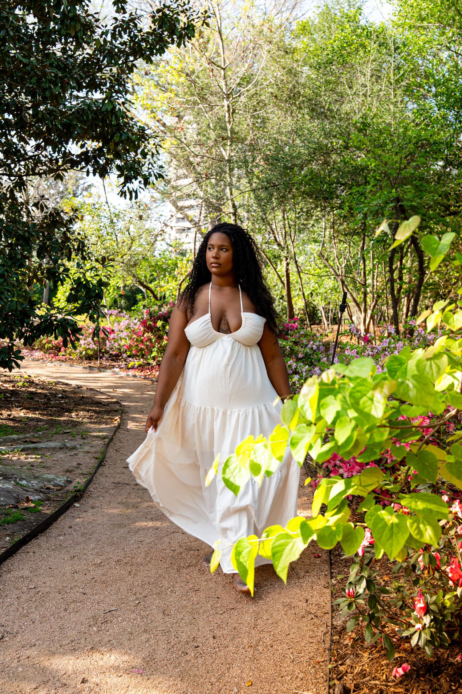 A Black woman in a flowing white maxi dress walks along a garden path surrounded by lush greenery and colorful flowers.