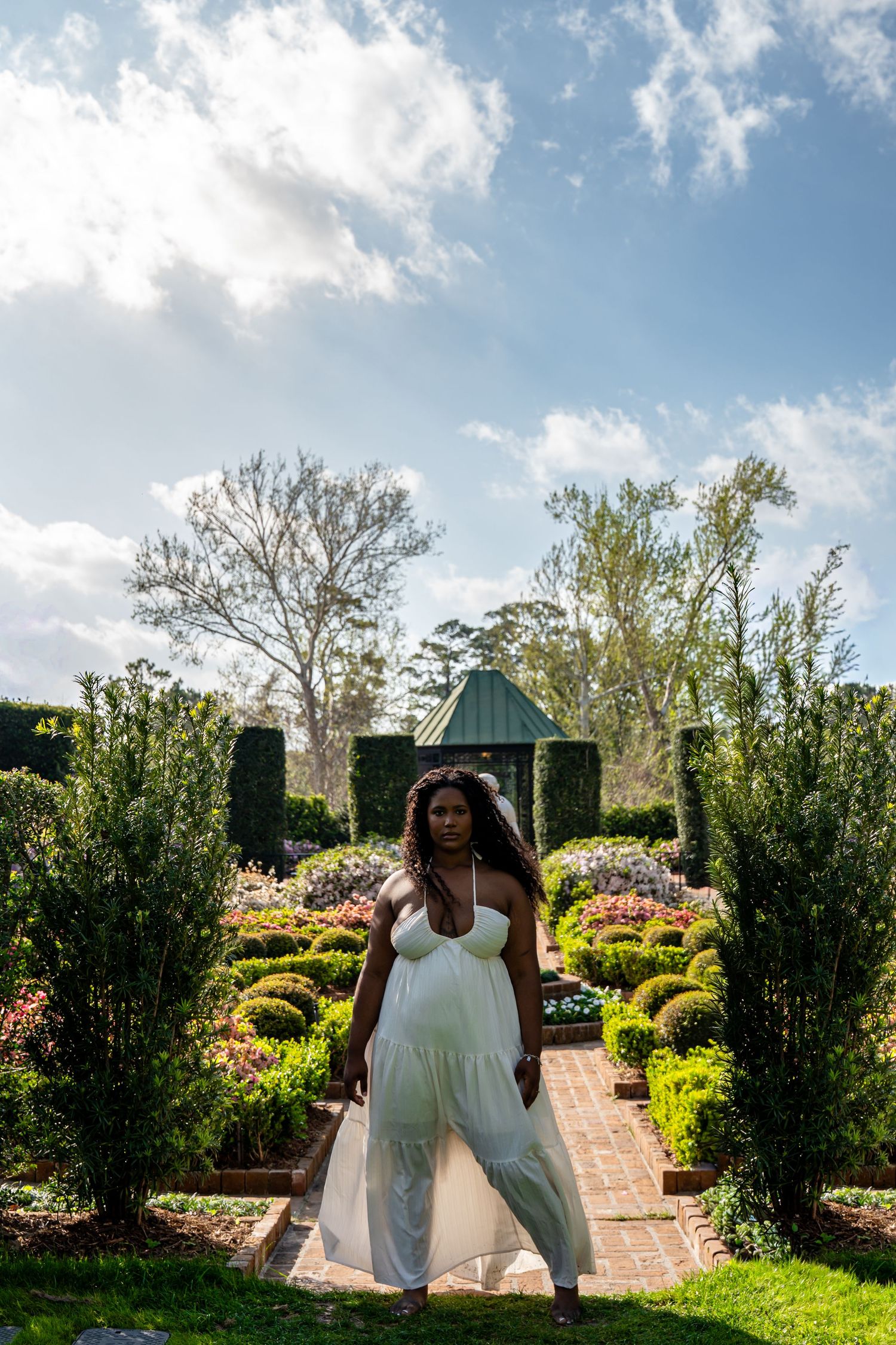 A Black woman in a white halter maxi dress poses in a formal garden with trimmed hedges and a green gazebo under blue skies.