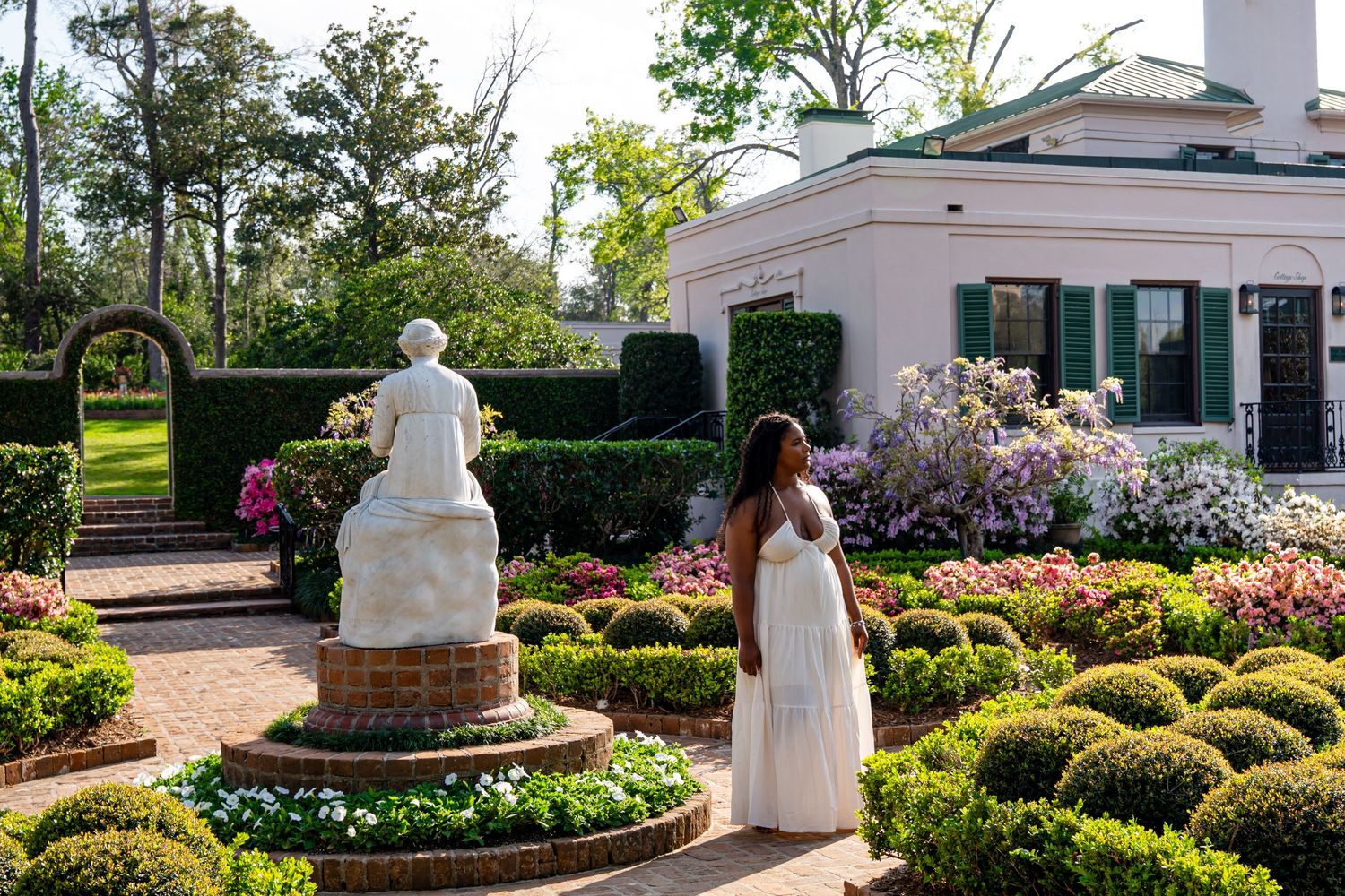 A woman in a white dress admires a classical statue centerpiece in a formal garden with manicured hedges and blooming flowers.
