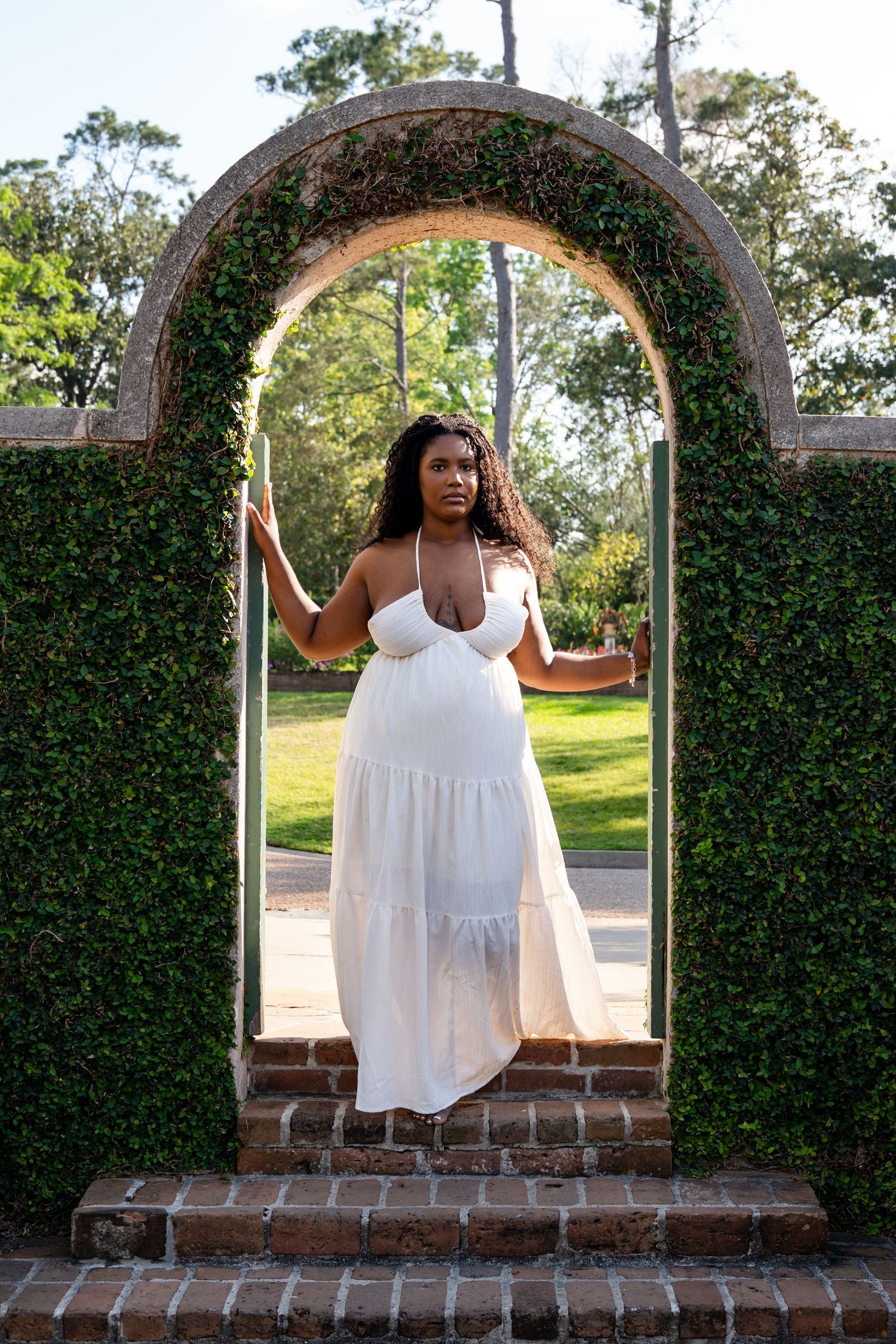 A Black woman in a white halter dress stands gracefully in a brick archway covered with lush green ivy in a garden.