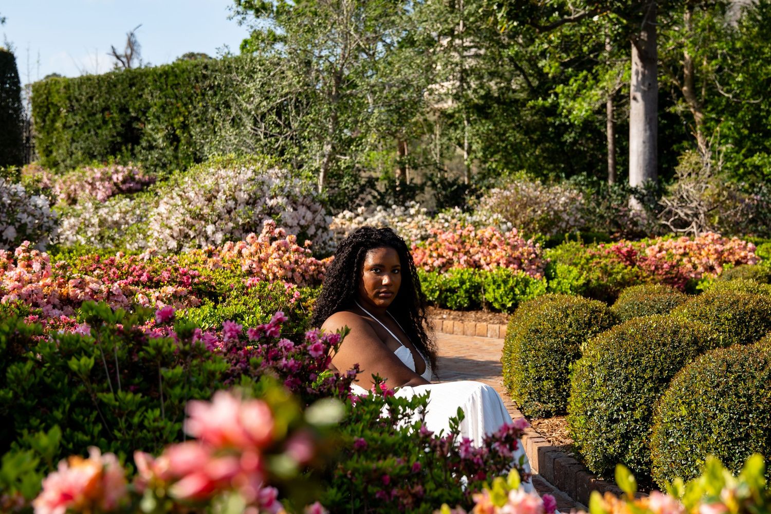 A Black woman sits among vibrant pink and coral azalea blooms and manicured boxwood hedges in a formal garden setting.