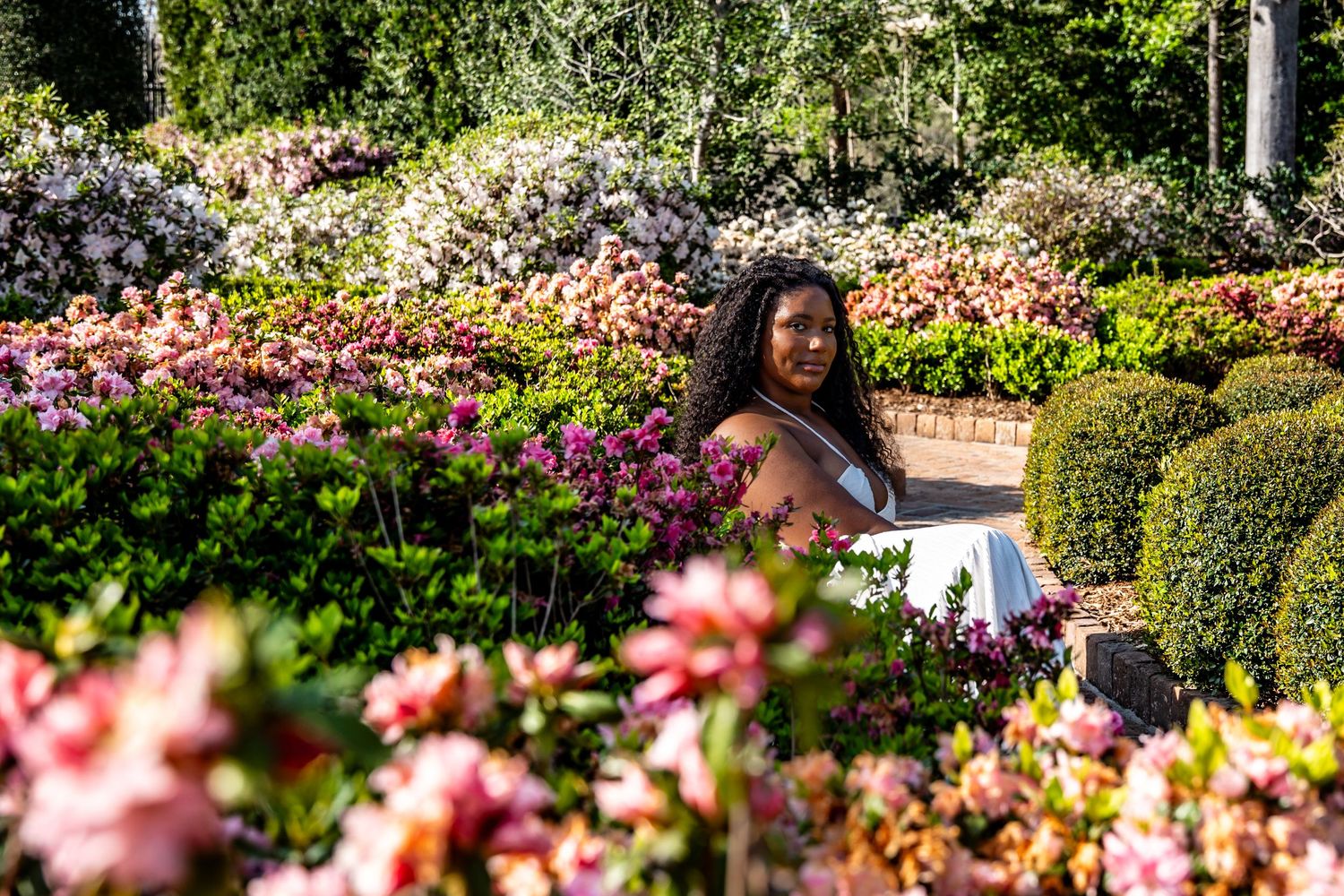A Black woman sits surrounded by colorful pink azaleas and neatly trimmed boxwood hedges in a formal garden landscape.