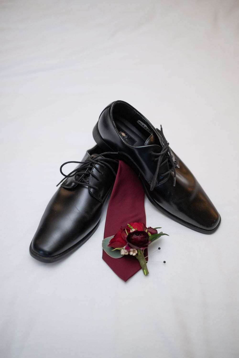 Black dress shoes and burgundy tie with boutonniere arranged on white background.