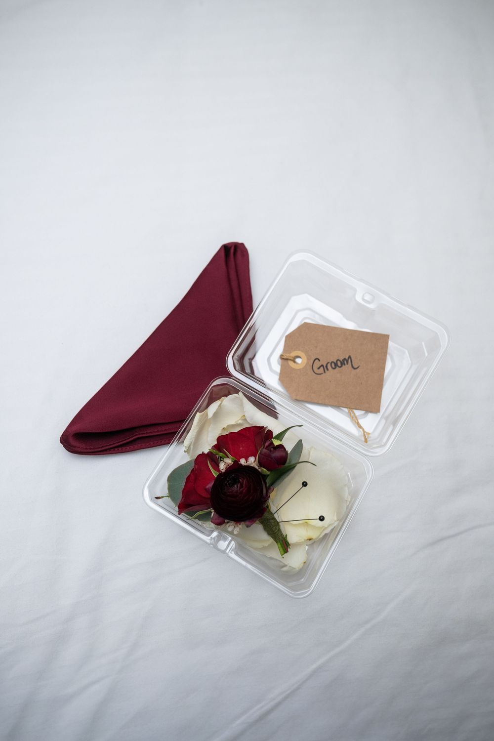 A burgundy napkin and red rose boutonniere with white accents are arranged in a clear plastic box.