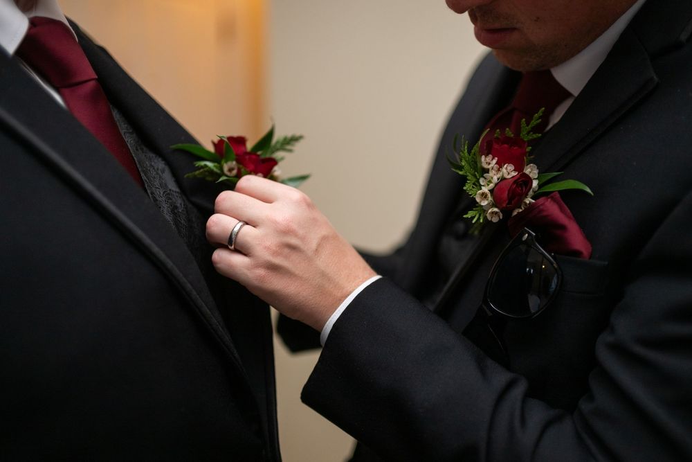 Close-up details of a groom's burgundy boutonniere and black suit jacket.