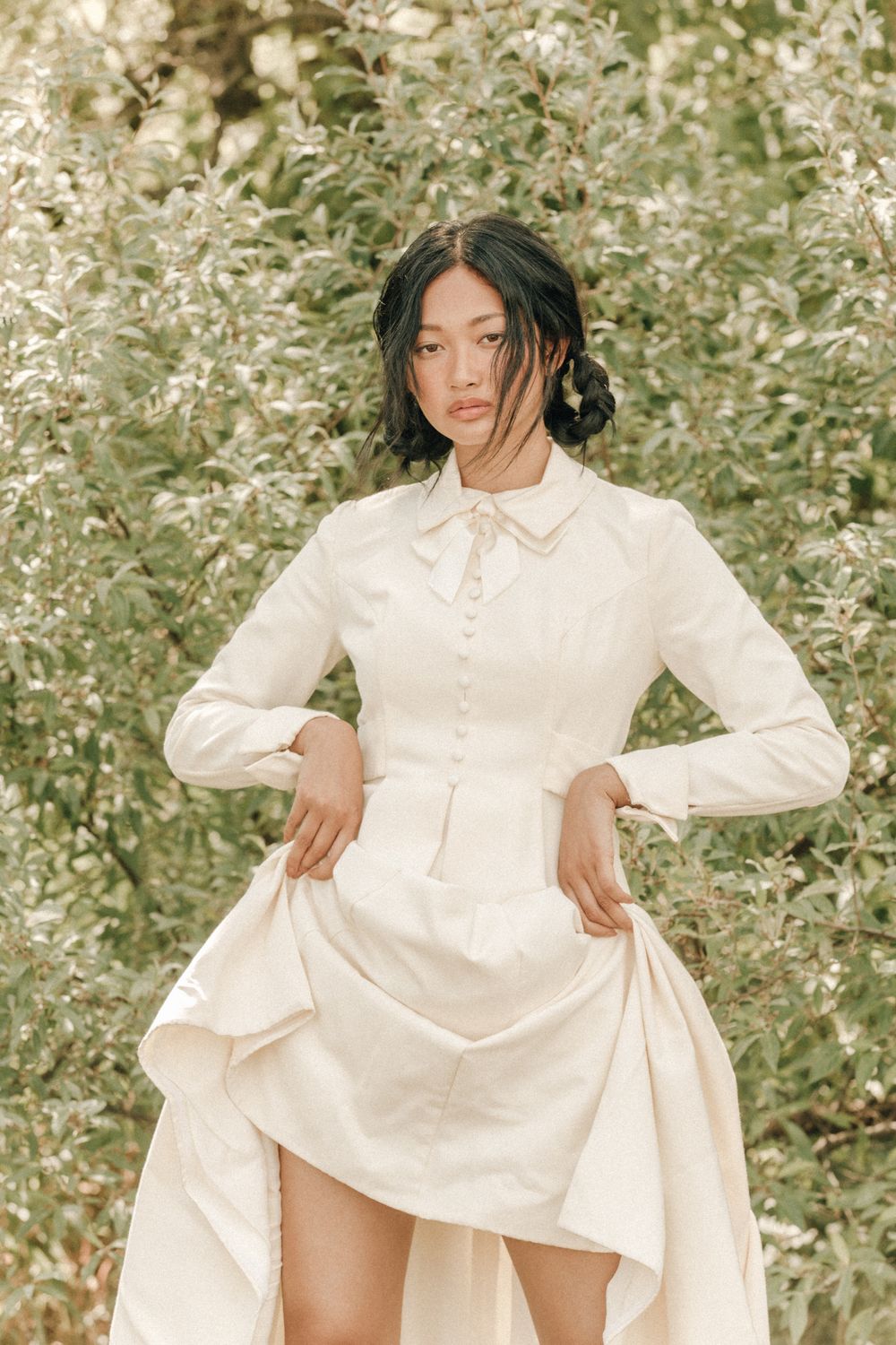 Fashion model poses in flowing white dress against blooming floral backdrop in ethereal outdoor photoshoot series.