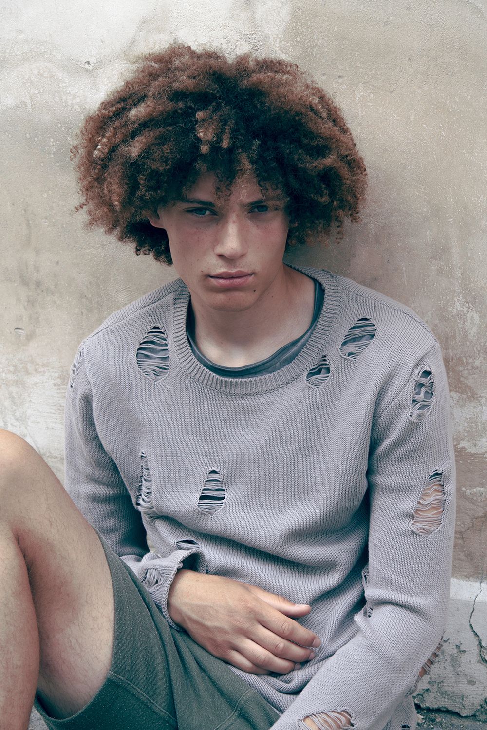 A person in a gray sweater with curly hair poses against a light colored wall.