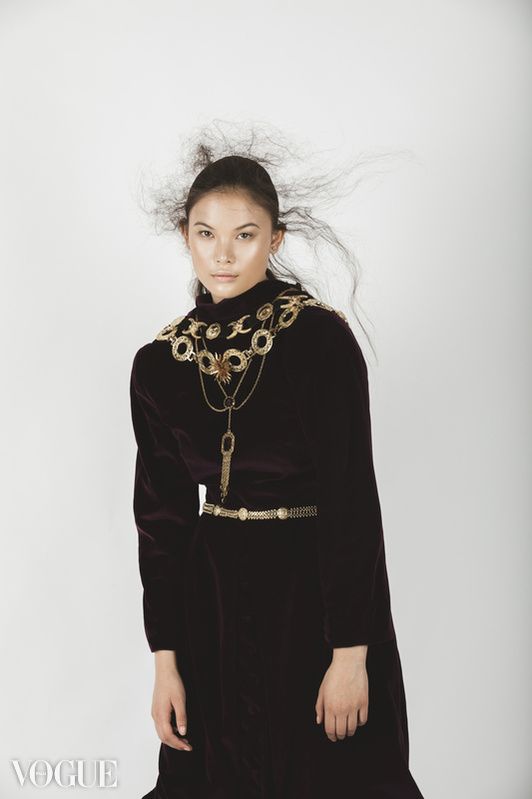 Fashion model wearing black velvet dress with ornate gold metallic necklace and belt against white background.