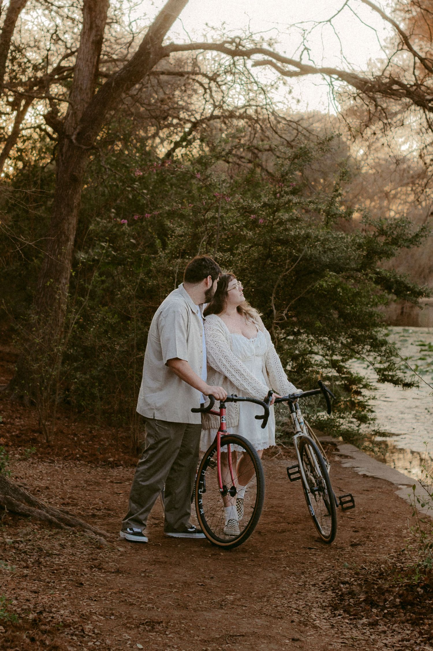 A couple shares a tender moment on a wooded path near a lake with a wheelchair visible in the scene.