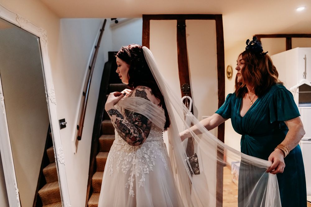 Someone helps with final adjustments to a white lace wedding dress with delicate details in a rustic interior setting.