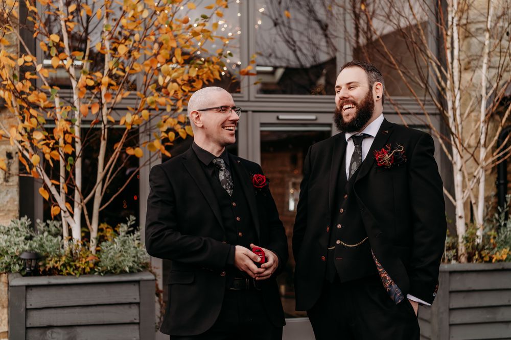 Wedding party members in black suits gather outdoors during autumn with golden foliage in the background.