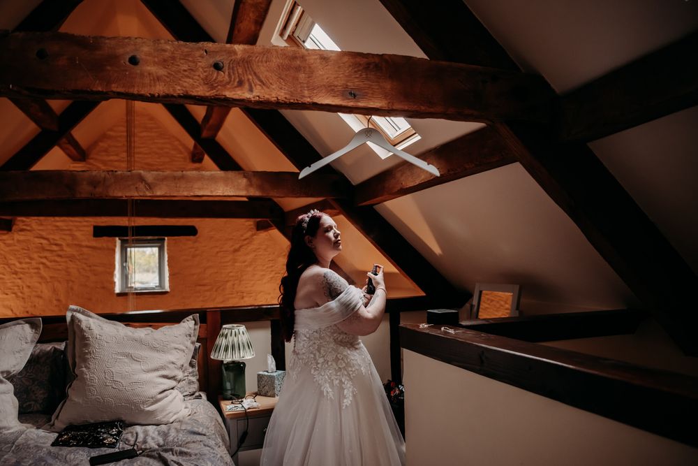 A bride in a white lace dress stands in a rustic attic room with exposed wooden beams and soft lighting.