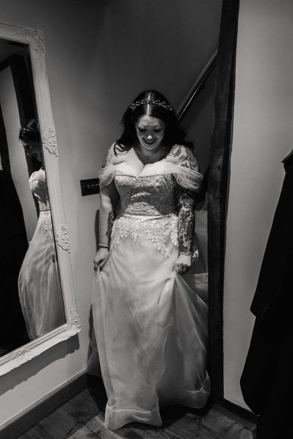 A bride in a white wedding gown stands by an ornate mirror in a black and white photograph.