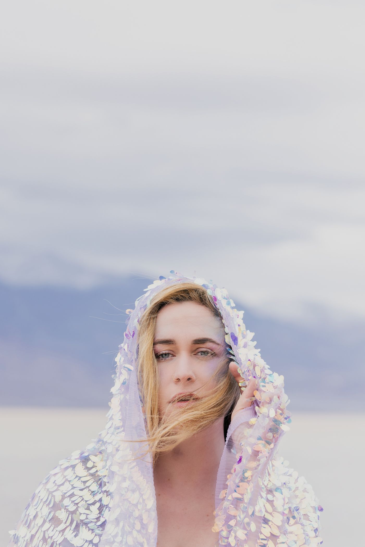 Artistic fashion portrait with shimmering holographic material draped against cloudy sky.