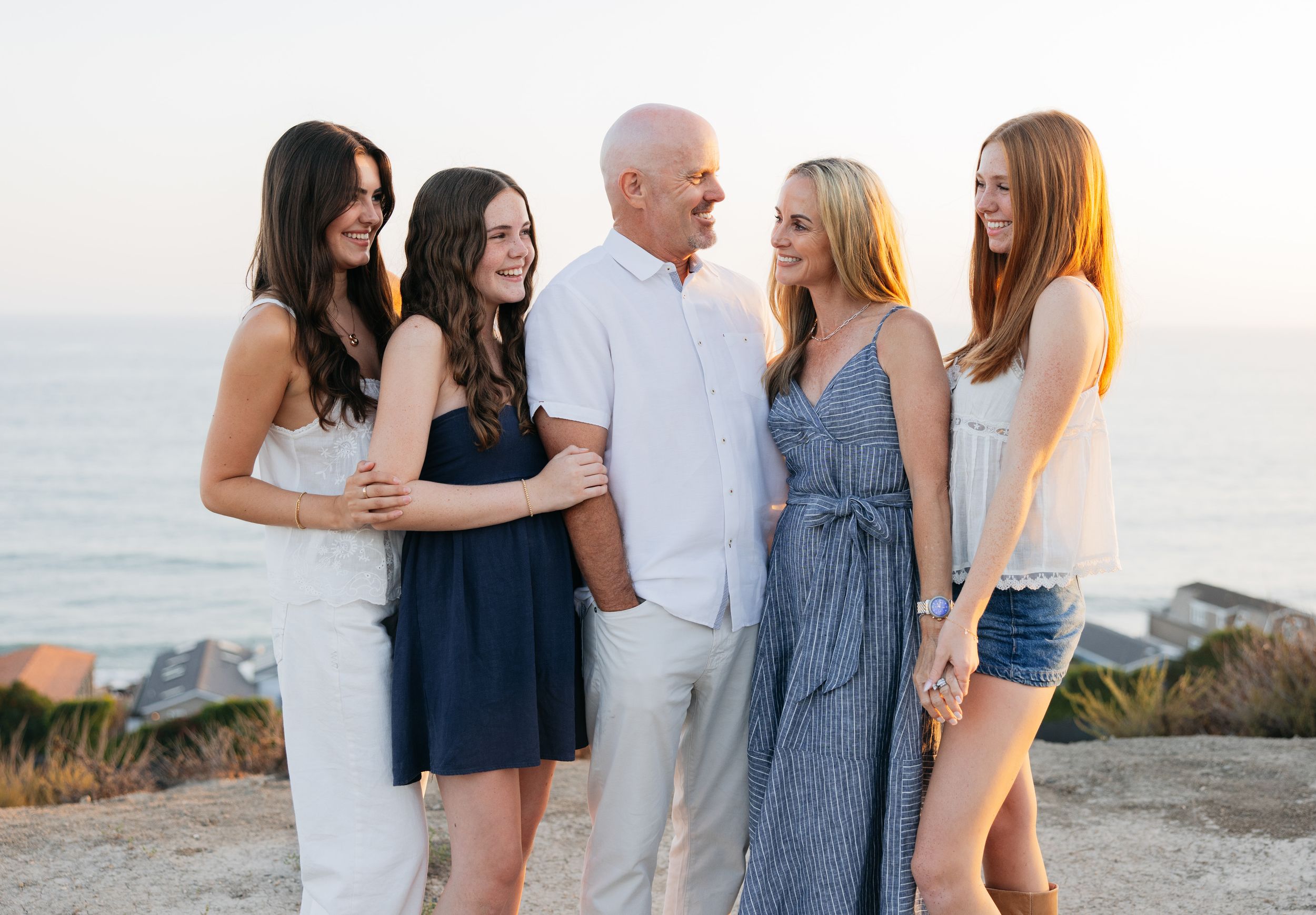 A group stands together on a coastal cliff during sunset, wearing casual summer outfits in blue and white tones.
