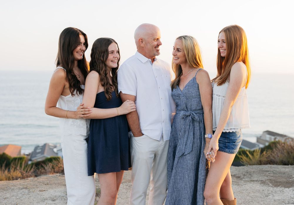 A group stands together on a coastal cliff during sunset, wearing casual summer outfits in blue and white tones.