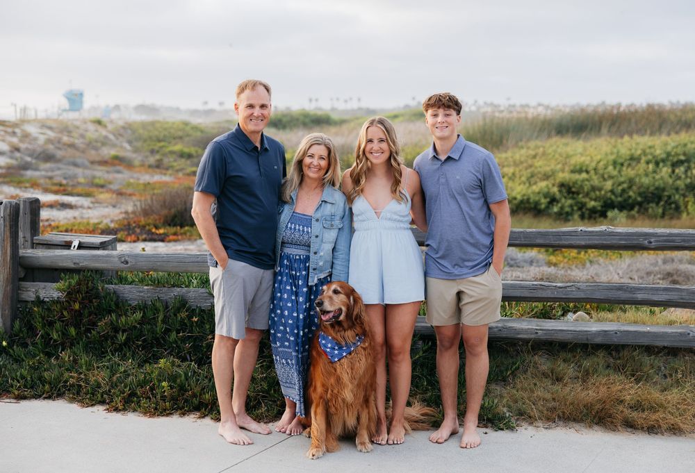 A group of people and their golden retriever pose together on a beach boardwalk at sunset.