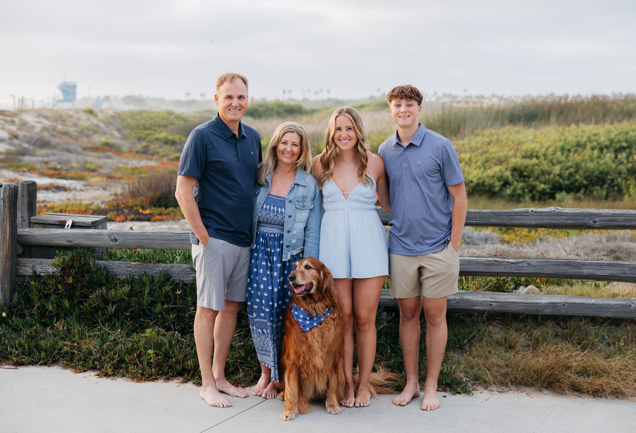 A group of people and their golden retriever pose together on a beach boardwalk at sunset.