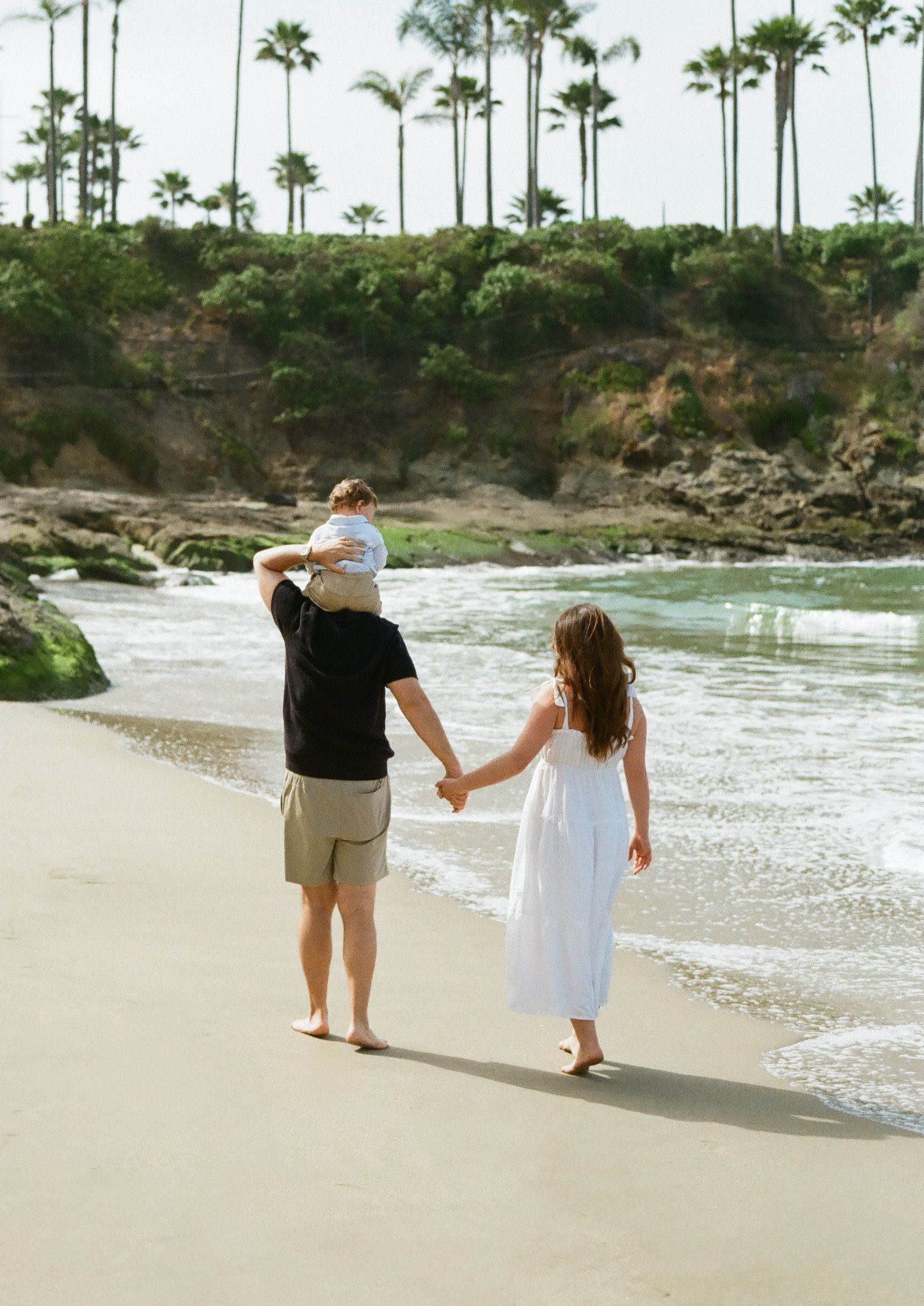 A couple walking hand in hand along a scenic beach with palm trees and cliffs in the background.