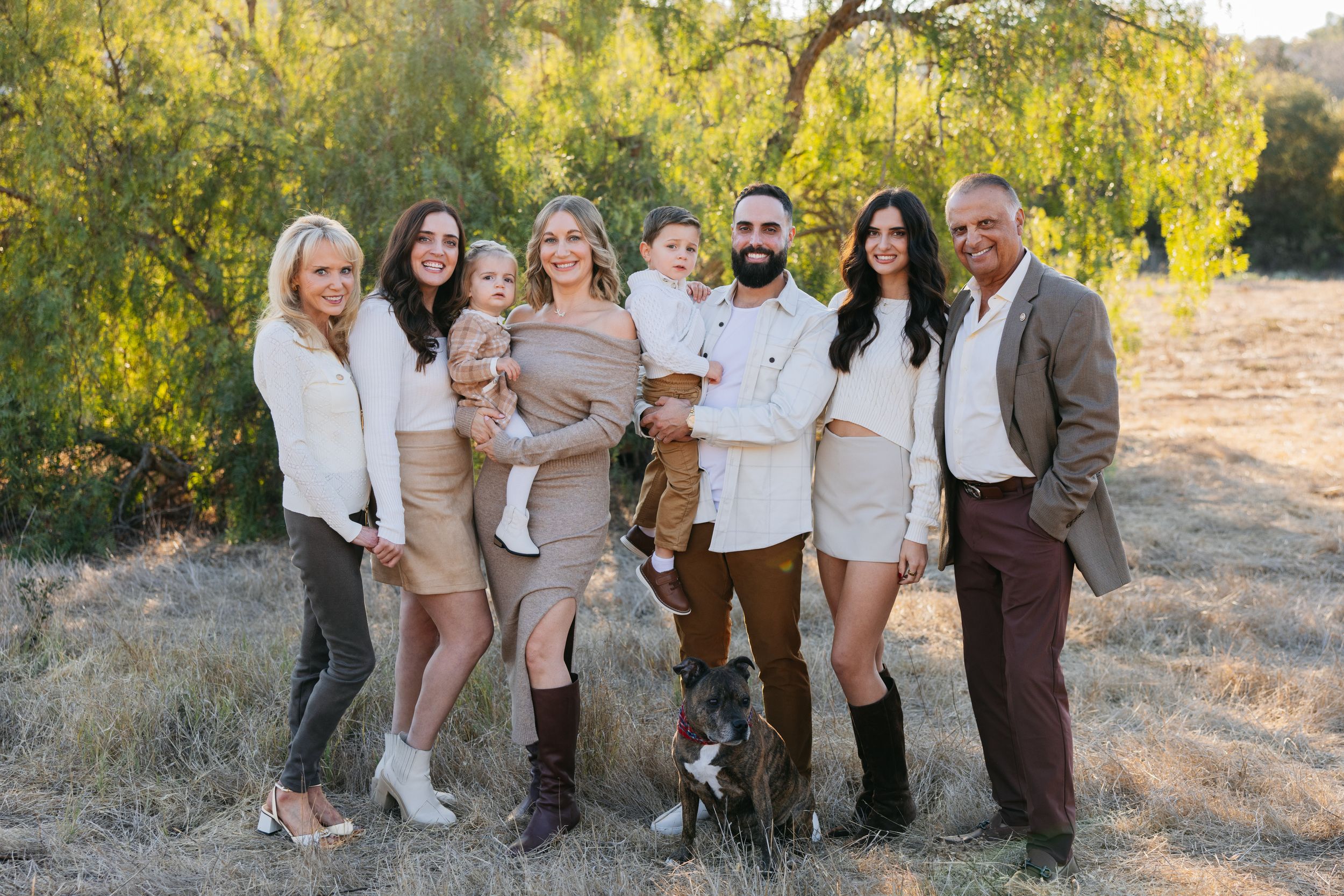 A group in coordinated cream and brown outfits pose together for outdoor family photos in a sunlit field.