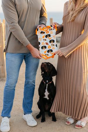 A woman in a brown dress and a person in jeans hold an orange cat-themed pillow while standing beside a black dog.