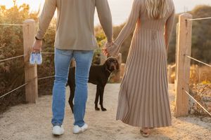 Couple walking their black dog on a trail at sunset while holding hands.