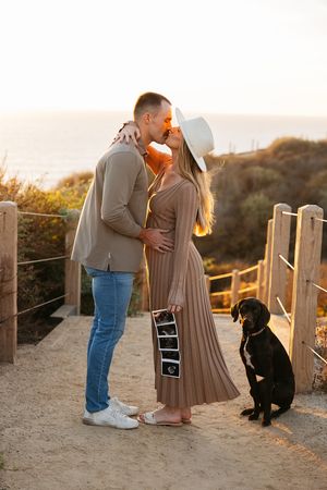 A couple shares a romantic moment on a beachside boardwalk at sunset with their black dog by their side.