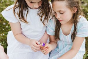 Two children in light dresses sit together in a field of daisies while examining a small purple flower.