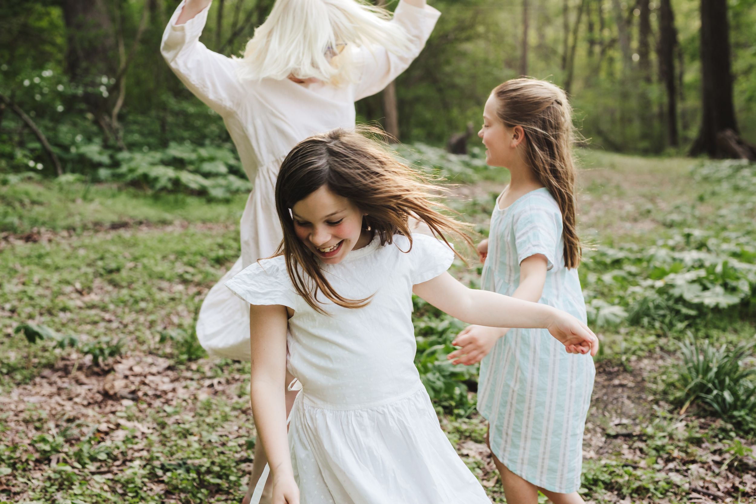 Three people in white dresses dance joyfully together in a lush green forest setting during springtime.
