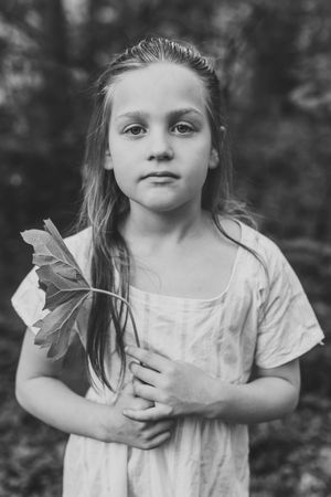 Black and white portrait of a young child holding a maple leaf while wearing a light-colored shirt outdoors.