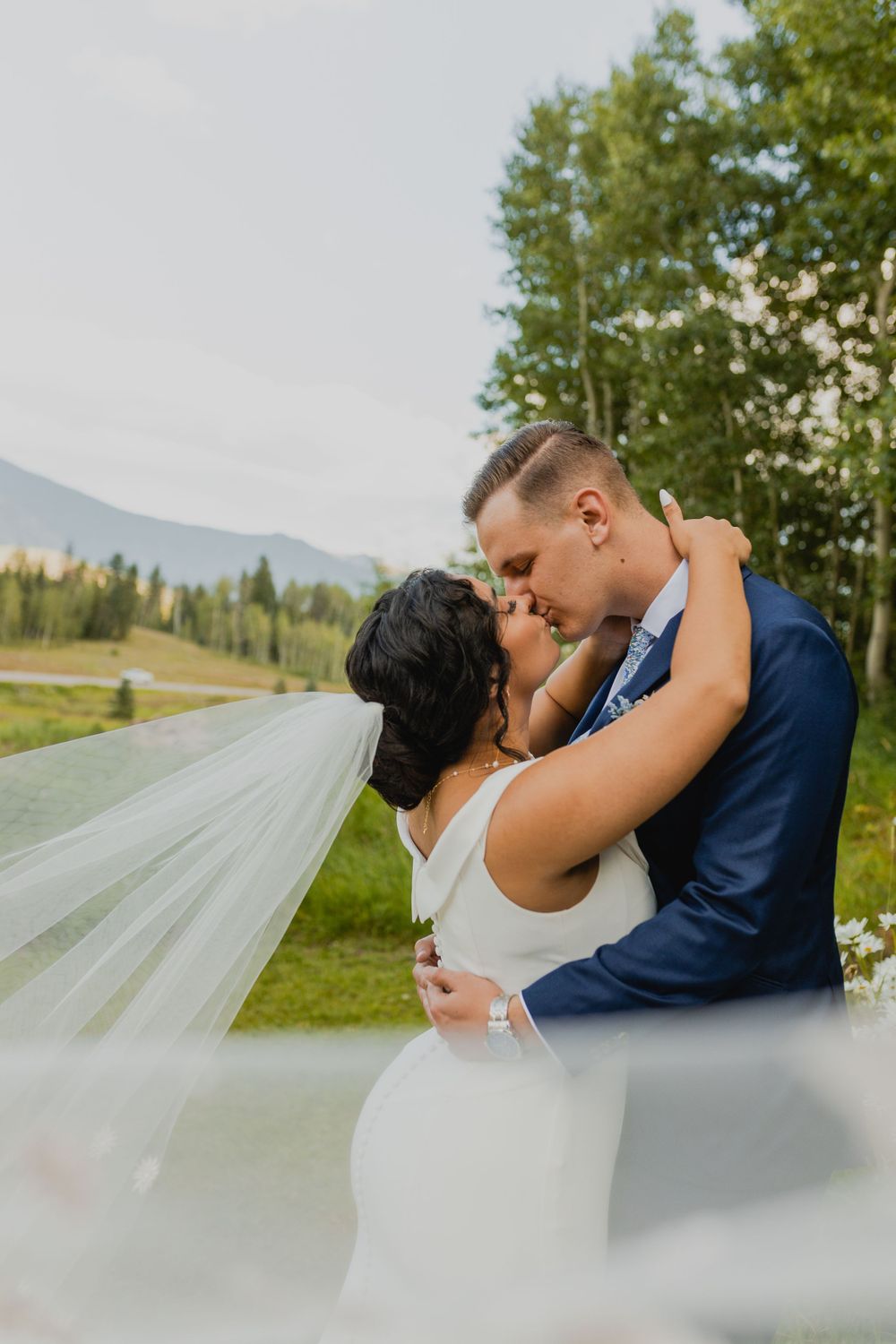 A romantic kiss captured against mountain scenery with flowing bridal veil.