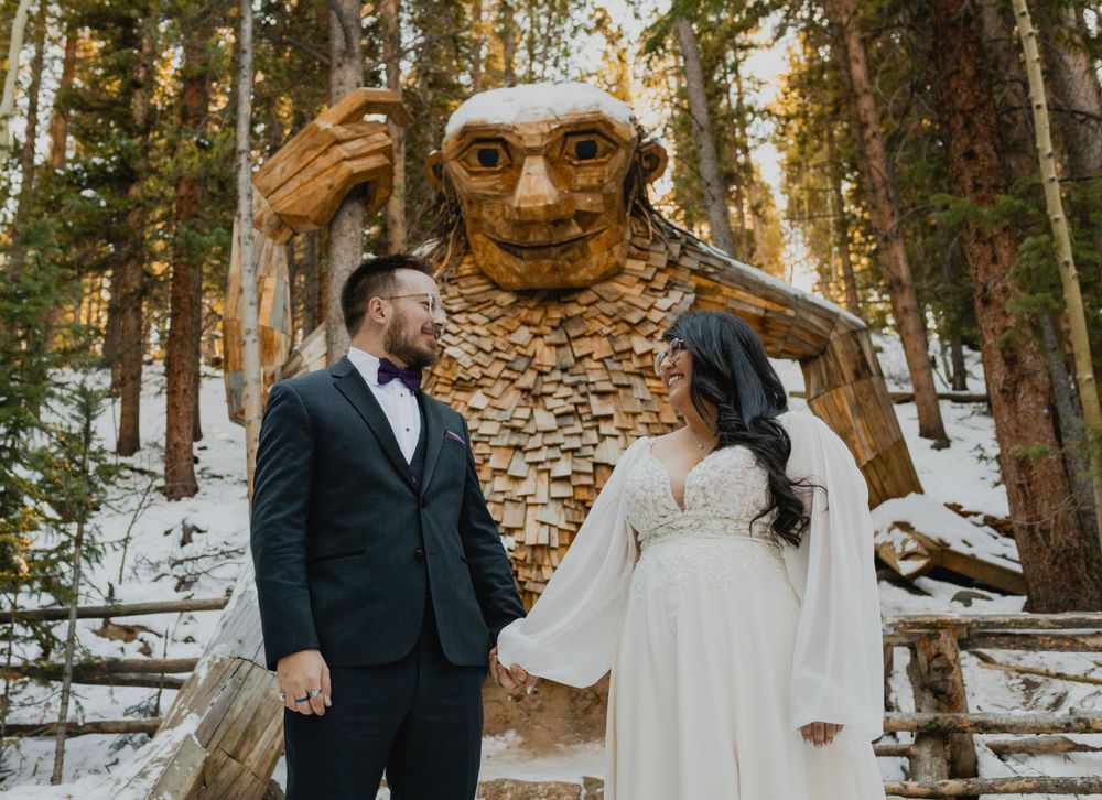 Winter engagement photo with quirky wooden troll sculpture in snowy forest setting.