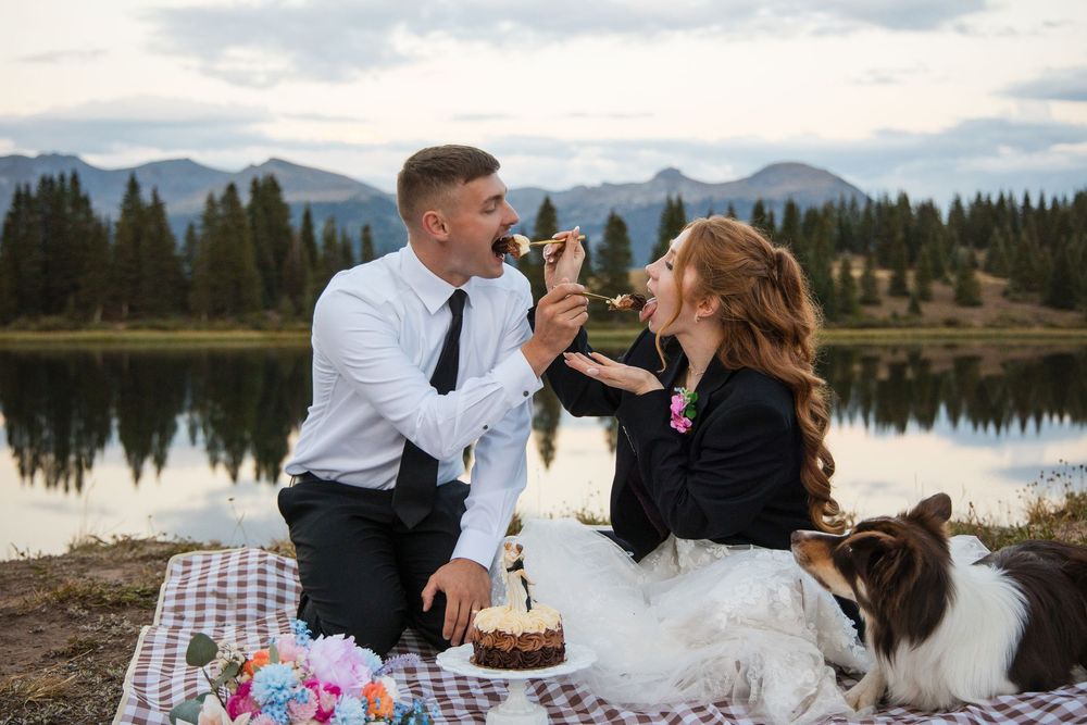 A couple enjoys a lakeside picnic with their dog in front of mountain views and evergreen trees.