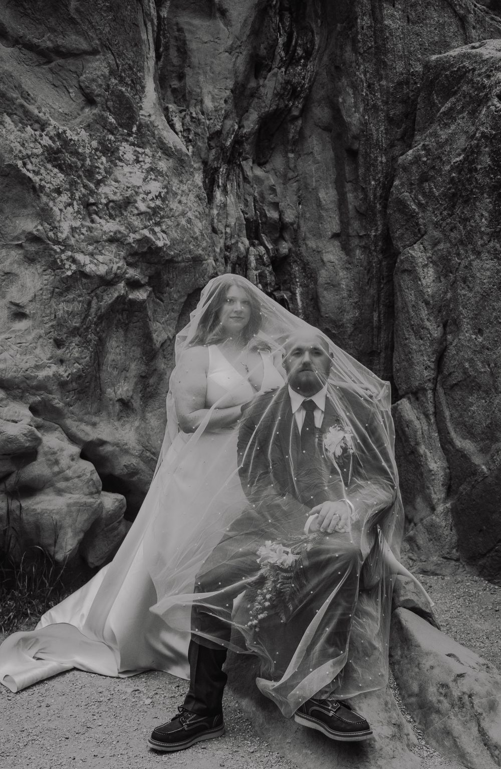 A black and white wedding photo of a couple covered in a veil against dramatic rocky cliffs.