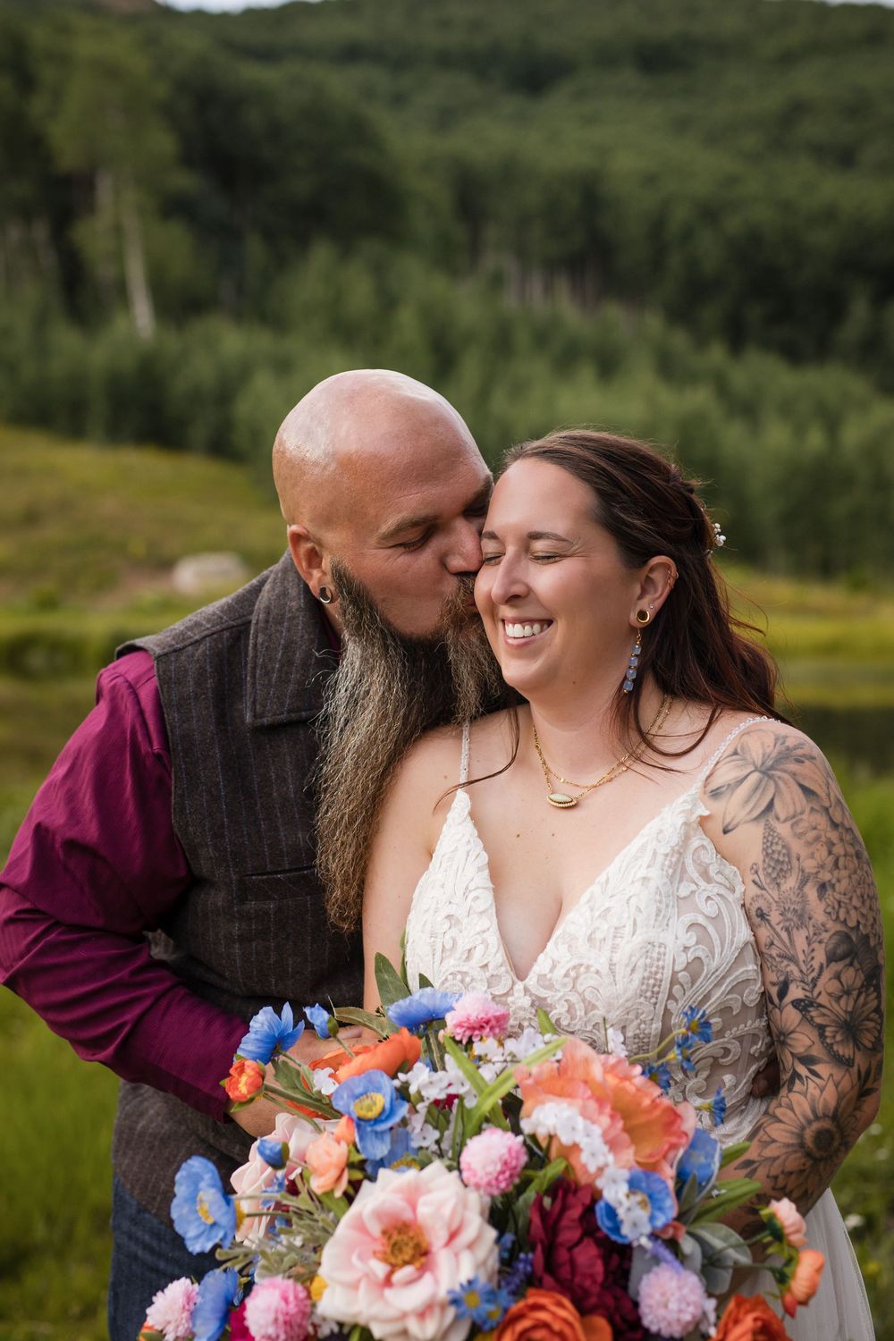 A joyful couple shares intimate moments in nature while holding a vibrant bouquet of pink, orange and blue flowers.