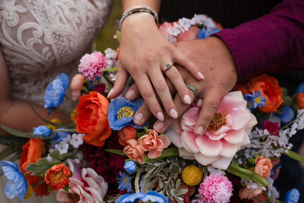 Hands displaying wedding rings rest on a vibrant bouquet of pink, orange, and blue flowers with lace in background.
