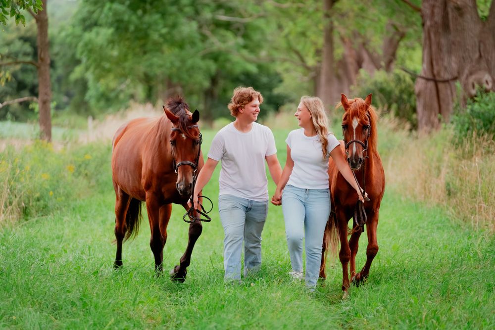 Couple walking two horses together on wooded trail.