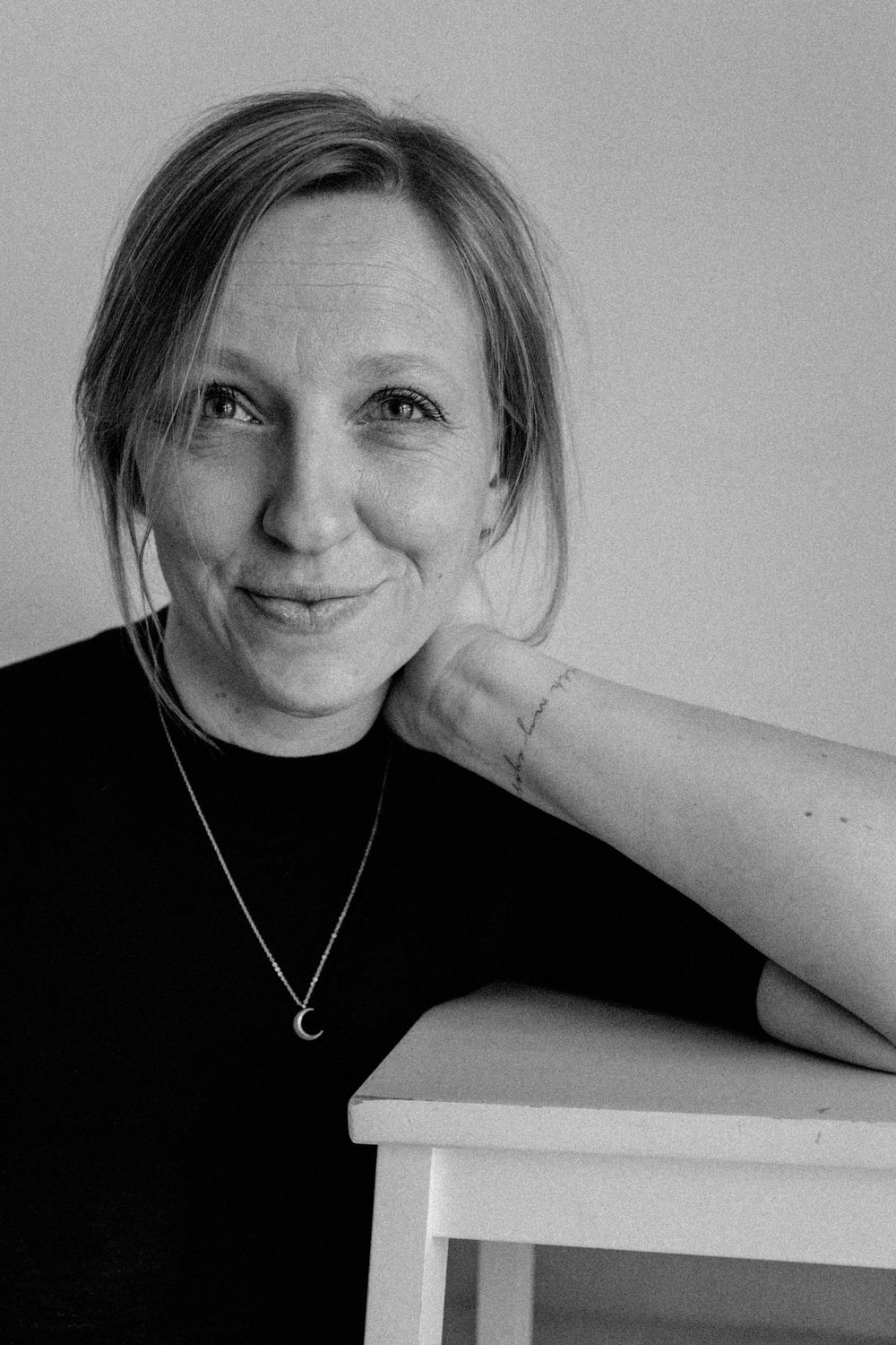 Black and white portrait of someone wearing a black sweater and crescent necklace, resting against a light surface.