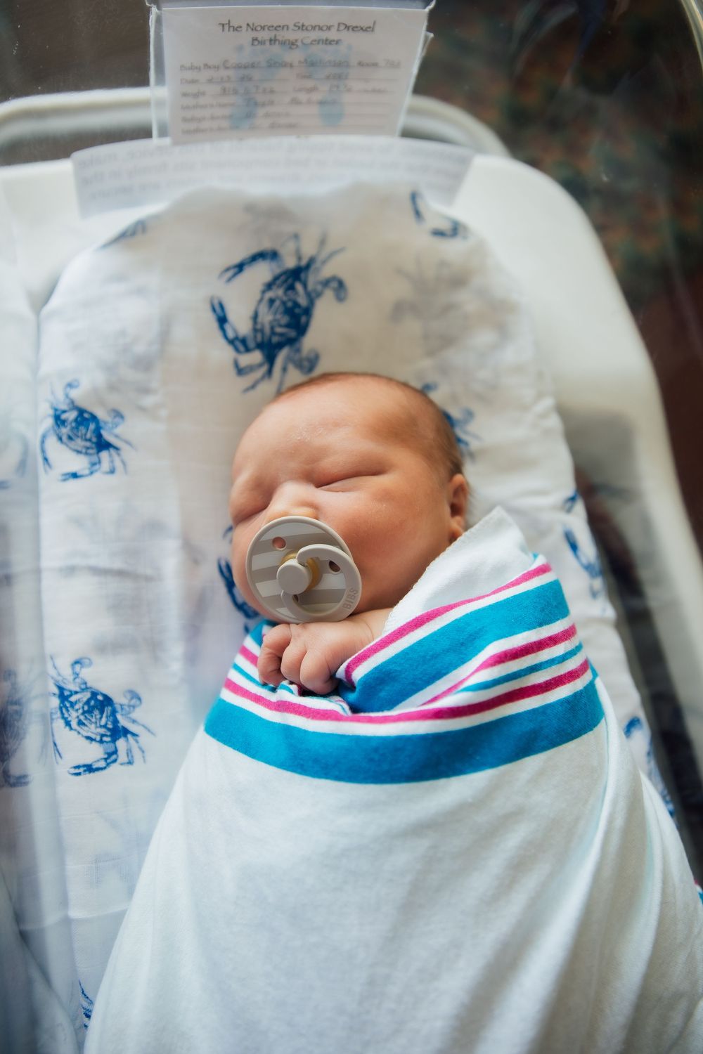 Newborn baby sleeping peacefully in hospital bassinet, swaddled in striped blanket with crab print, using a pacifier. Newport Hospital fresh