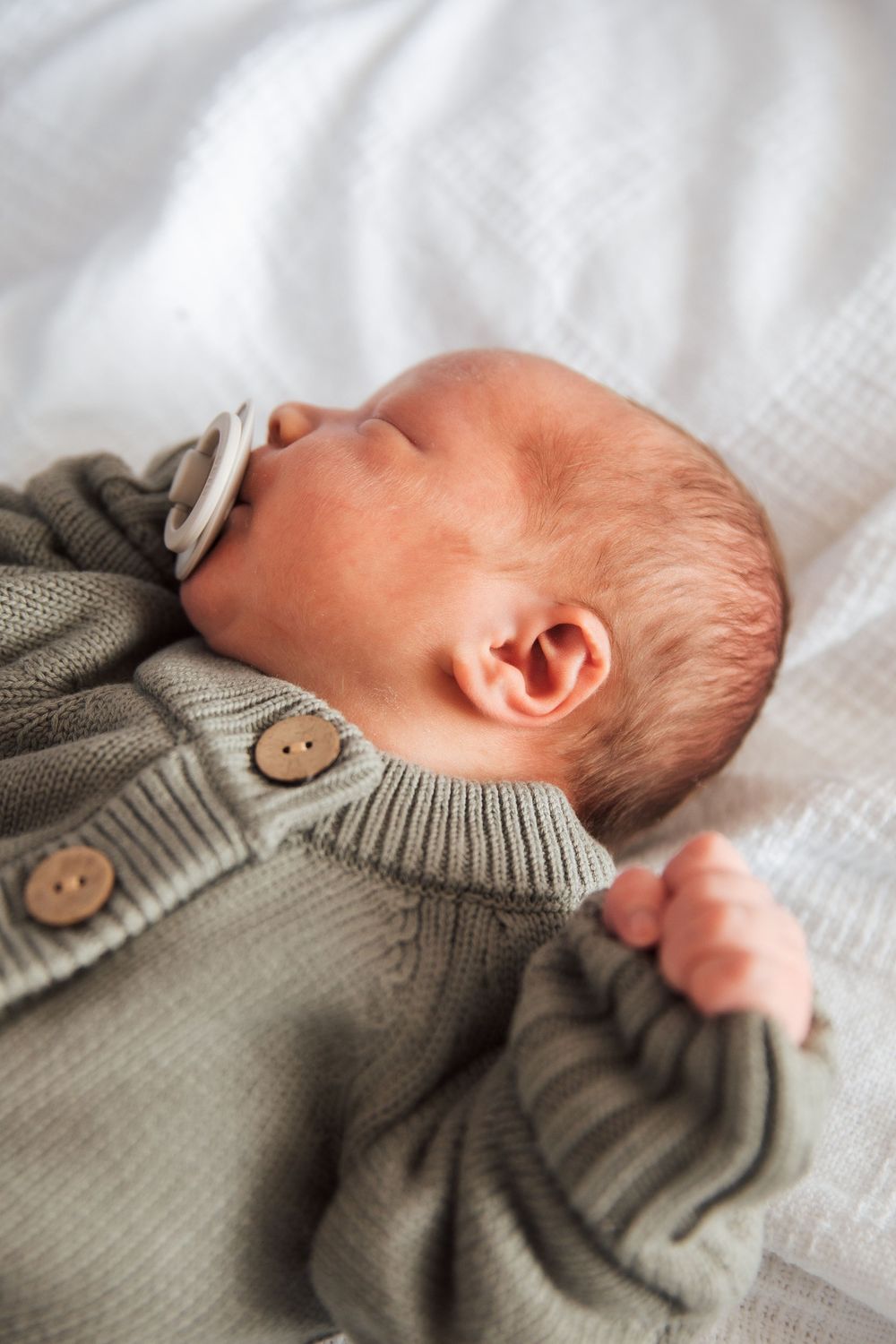 Newborn baby sleeping peacefully in a sage green knit sweater with buttons, lying on a white blanket. Newport Hospital fresh 48