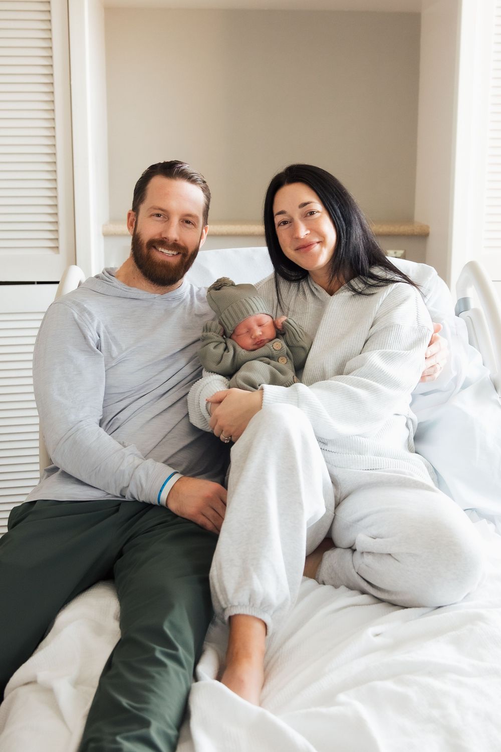 Happy couple sitting on a hospital bed smiling while holding their newborn baby dressed in a green outfit. Newport Hospital fresh 48