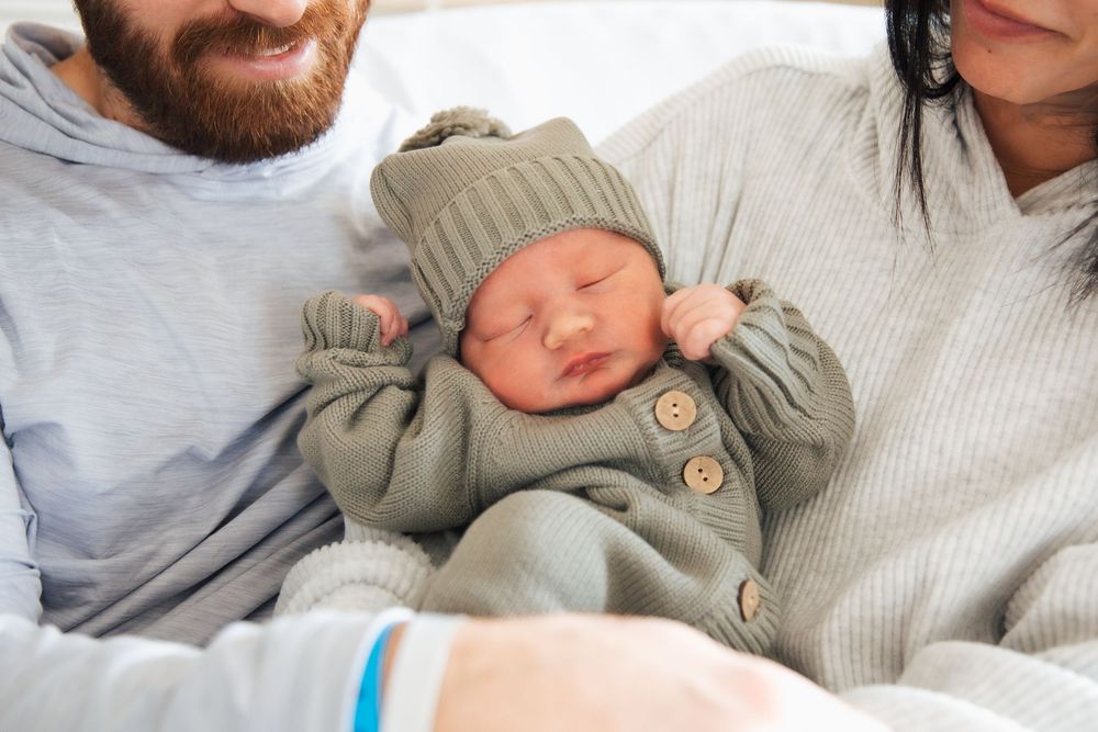 Newborn baby sleeping in sage green outfit and hat, cradled by parents in a cozy hospital setting. Newport Hospital fresh 48
