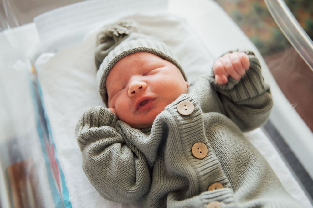 Newborn baby sleeping in hospital bassinet wearing gray knit cardigan and matching pom-pom hat. Newport Hospital fresh 48