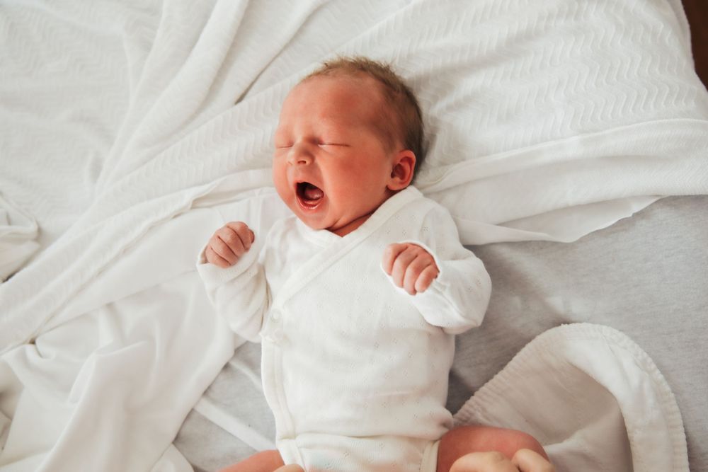 Newborn baby yawning while wearing white onesie on white bedding. fresh48. Newport hospital