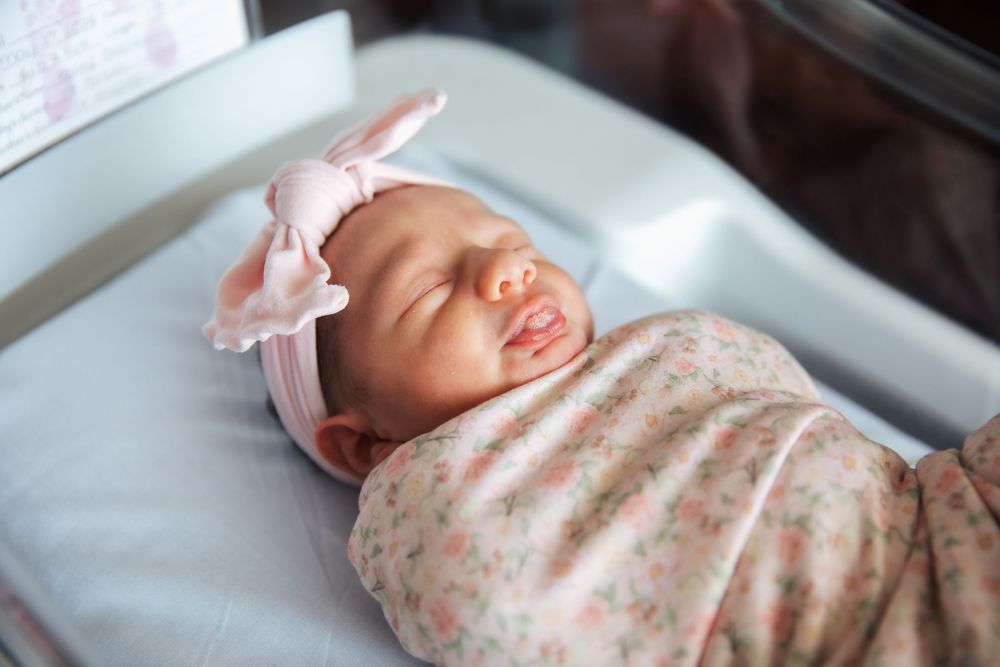 Newborn baby girl swaddled in floral blanket wearing pink bow headband sleeping peacefully in hospital bassinet. fresh 48, Newport Hospital